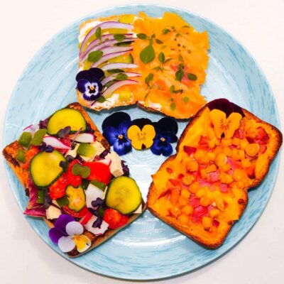 A top down view of 3 types of ciabatta sandwiches, one with salmon and chives, another one with cheese, ham and corn and another vegetable one, with tofu and edible flowers