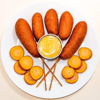 A top down view of some gluten free corn dogs, 4 of them on skewers and 10 small slices, served with mustard