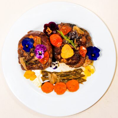 A top down view of a ready to be served lamb steak, topped with baked cherry tomatoes, asparagus, carrot disks and edible flowers